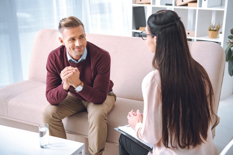 smiling patient sitting on couch and looking at psychotherapist with clipboard in office